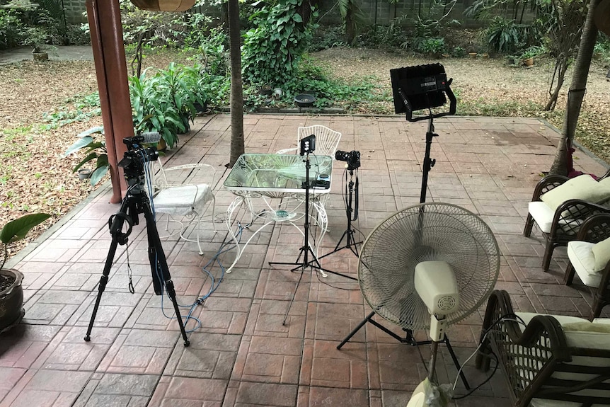 Cameras and lights on tripods pointed at two chairs next to glass table with garden background.