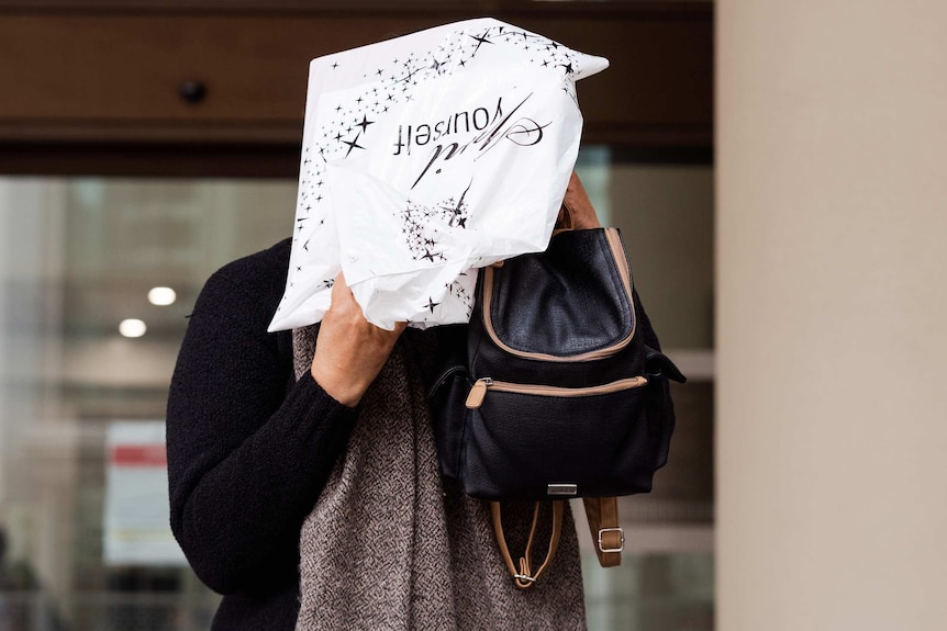 A woman holds a plastic bag and a backpack up to cover her face outside court.