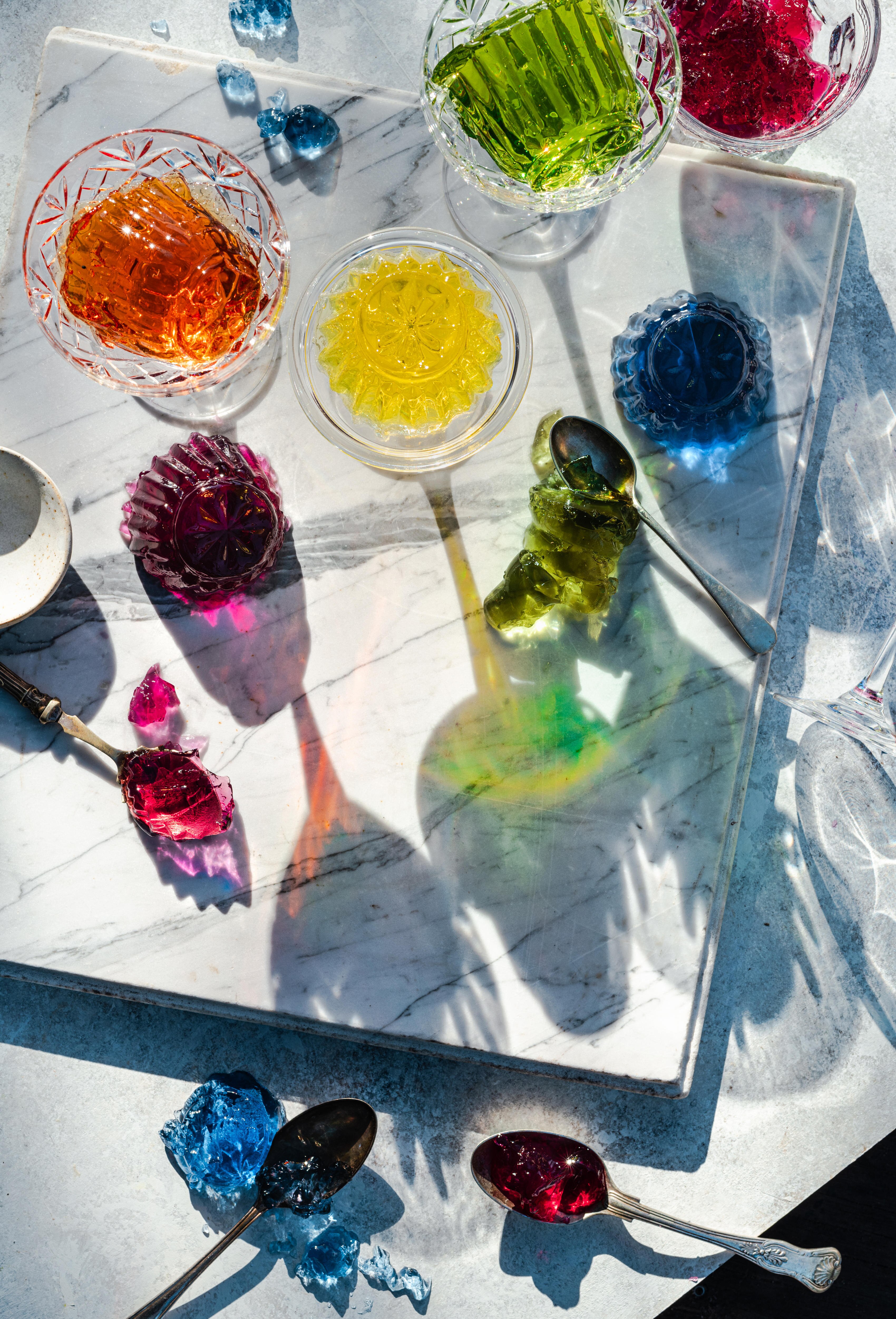 Glasses of different coloured jellies sit on a marble benchtop. Spoons sit amongst dropped blobs of jelly. 