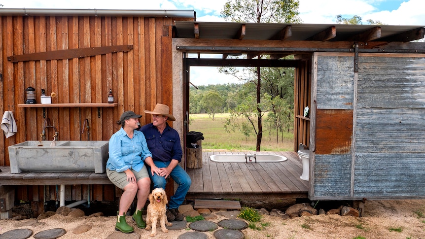 A man and woman sit embracing outside a rustic cabin.