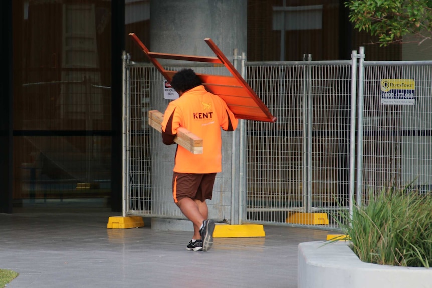 A man in an orange shirt carrying things into a building.