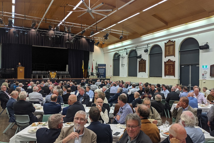 A group of 60-something men drink wine and eat dinner in an old school hall