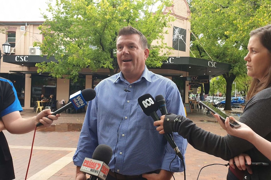 A man in a blue shirt stands facing media reporters with microphones