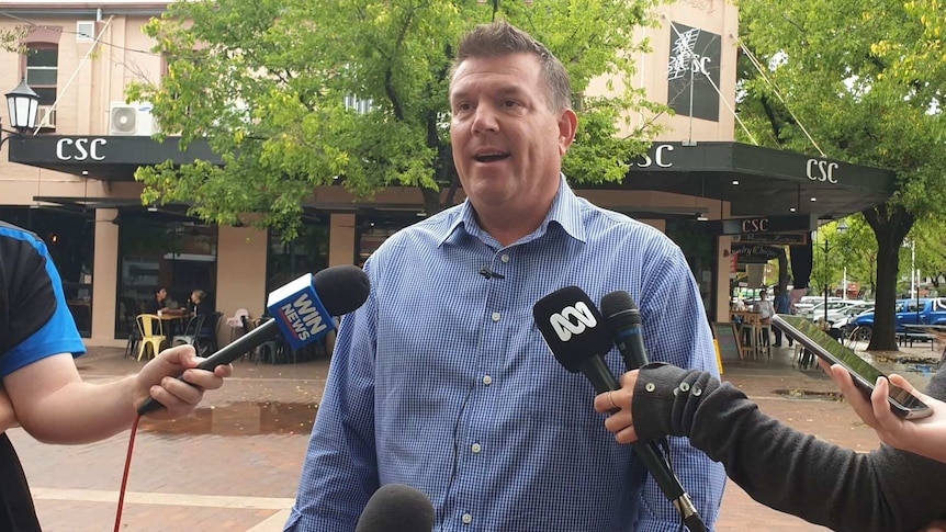 A man in a business shirt stands outside speaking to the media.