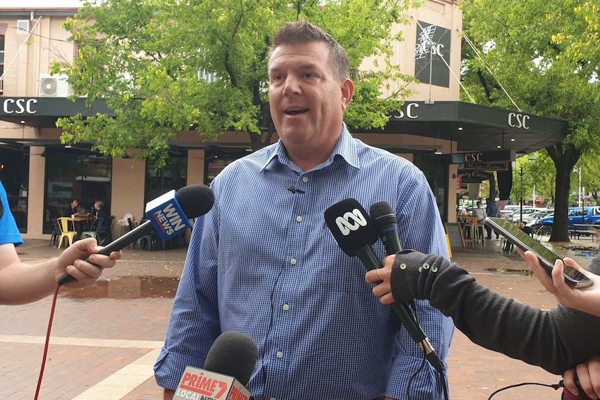 A man in a business shirt stands outside speaking to the media.