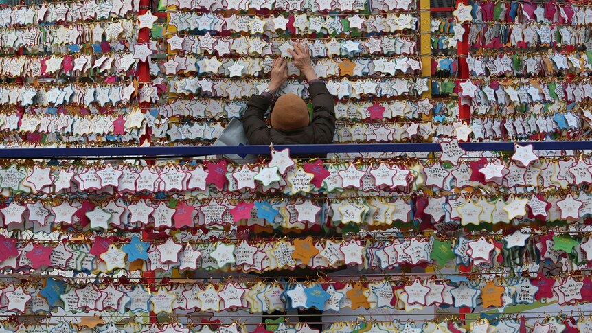 Rows and rows of star-shaped paper wishes line walls in a temple, with one person in the middle of the frame placing another one