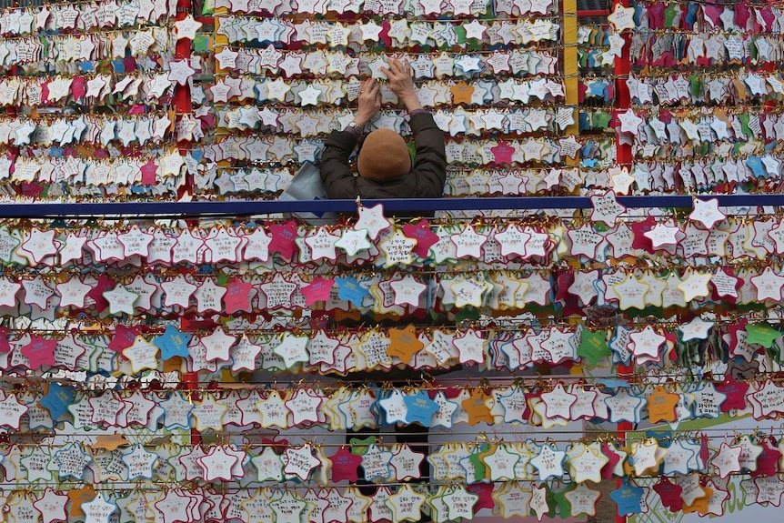 Rows and rows of star-shaped paper wishes line walls in a temple, with one person in the middle of the frame placing another one