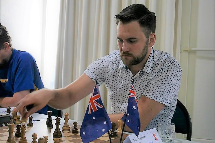 A man in a white shirt playing chess at a table with an australian and new zealand flag in view