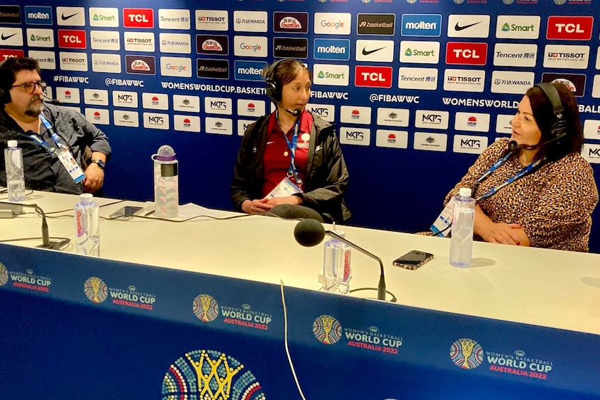 Three people with headphones sit at a table recording a podcast, with FIBA Women's Basketball World Cup signage behind them.