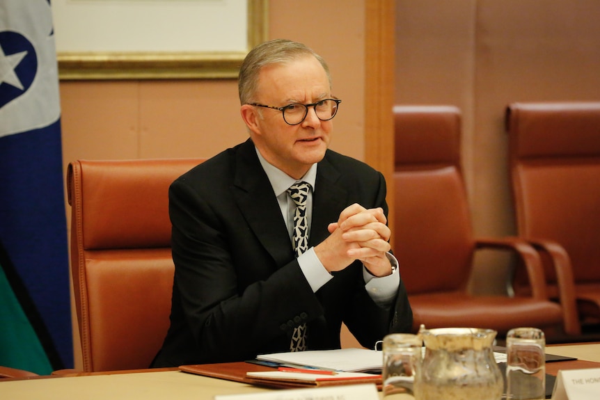 Albanese sits with his hands folded at a desk in a meeting room.