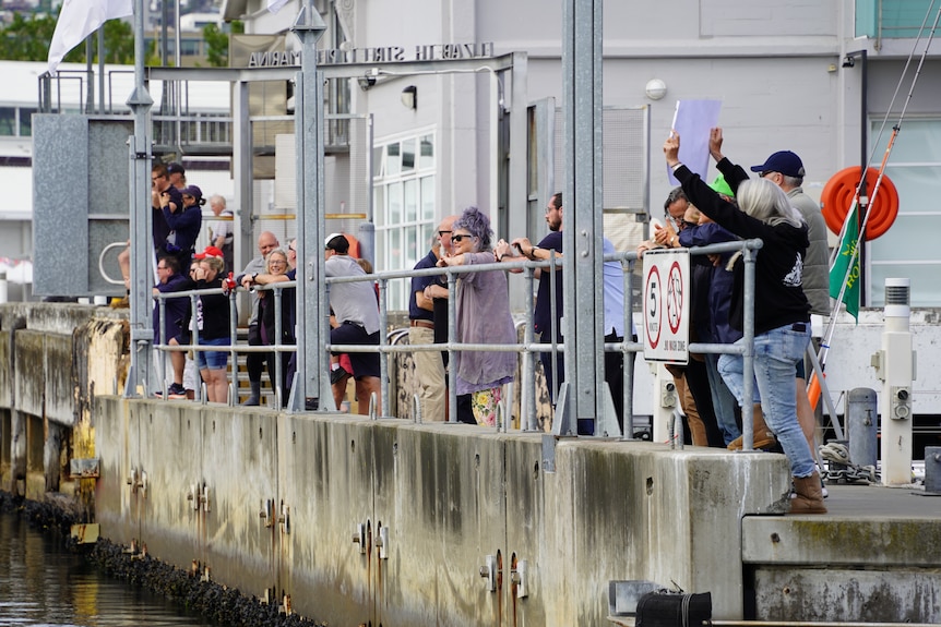 A line of people, some holding placards, greet a yacht.