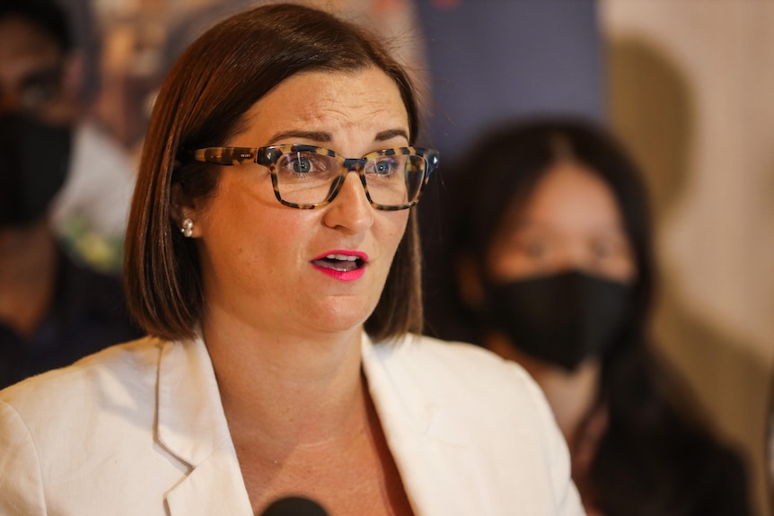 A woman in a white jacket and glasses speaks at a media conference. 