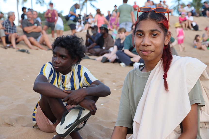 A young Indigenous woman sits on the beach, smiling at the camera, next to a young boy.