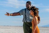 A tall Black man gestures to a short Black woman as they stand in a desert 