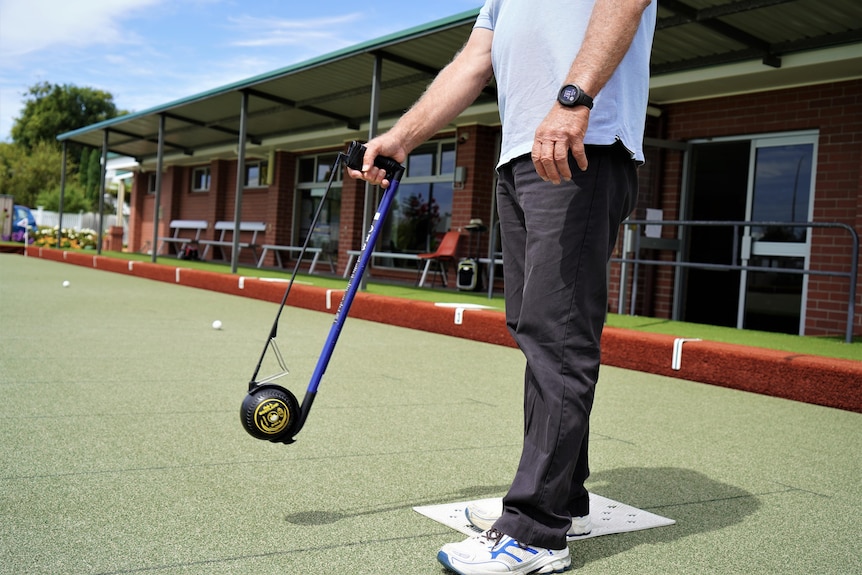 A man on a bowling green about to take a shot using an arm-extending device.