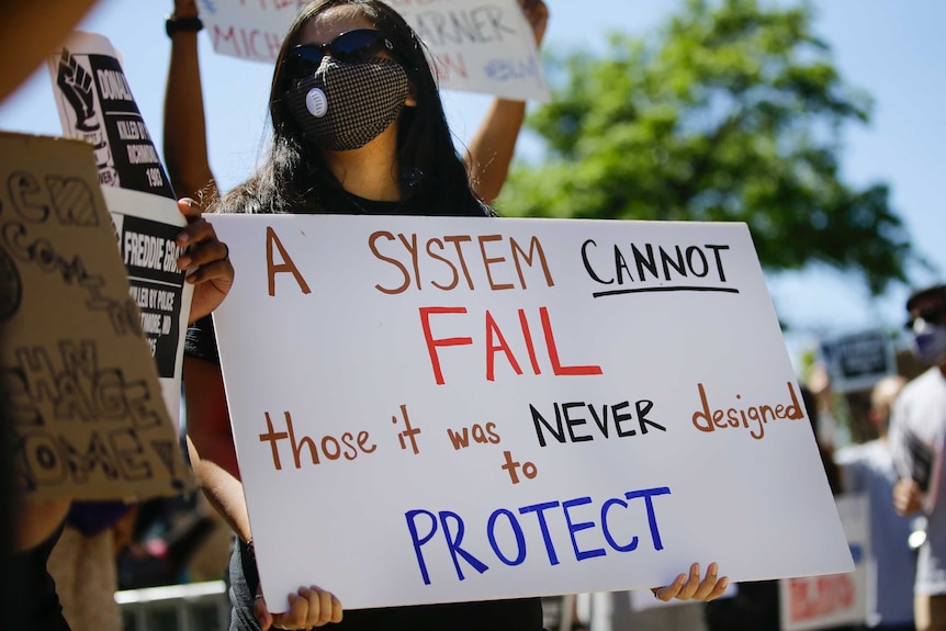 A young female in a black face mask and holding a white sign stands in a rally under blue skies.
