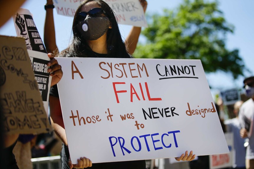 A young female in a black face mask and holding a white sign stands in a rally under blue skies.