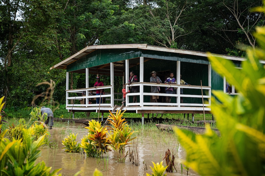 Un grupo de hombres y mujeres se encuentran en un porche cerca de un lago mientras una mujer recoge objetos del suelo.