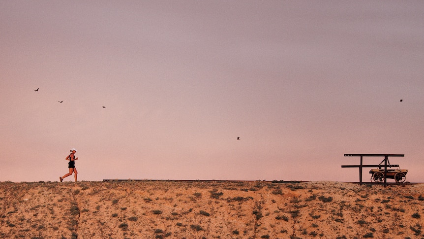 In the distance a woman runs across a dry landscape in front of a pink sky.