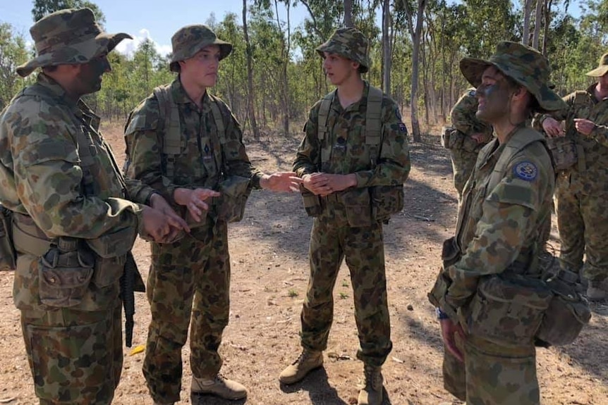 Four young men stand together having a chat in full cadet uniforms with plenty of gum trees behind.