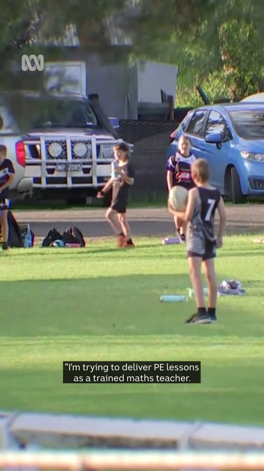 Three young people in footy garb stand on a grassy piece of land, the closest person holds a football
