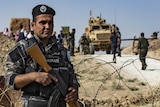 A man in military gear holding an AK-47 stands in front of barbed wire with other military equipment behind him.