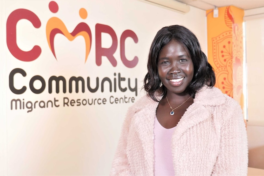 A Sudanese woman wearing a pink jacket and shirt smiles in front of a bright blue background.