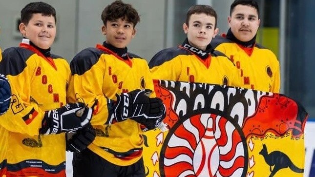 four indigenous male ice hockey players standing on the ice holding a flag wearing red and yellow uniforms