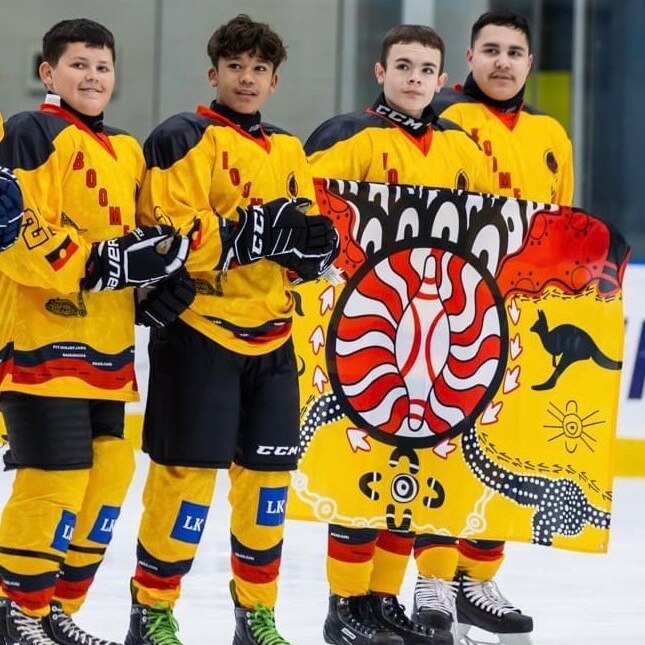 four indigenous male ice hockey players standing on the ice holding a flag wearing red and yellow uniforms