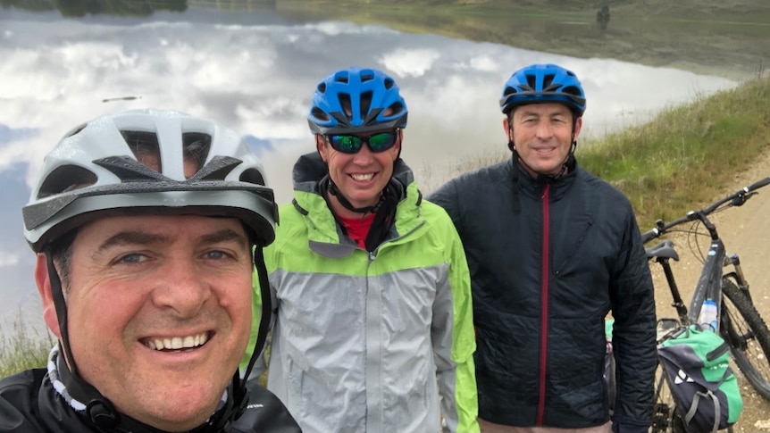 Three men in bike helmets smile to the camera, with a bike also in shot.