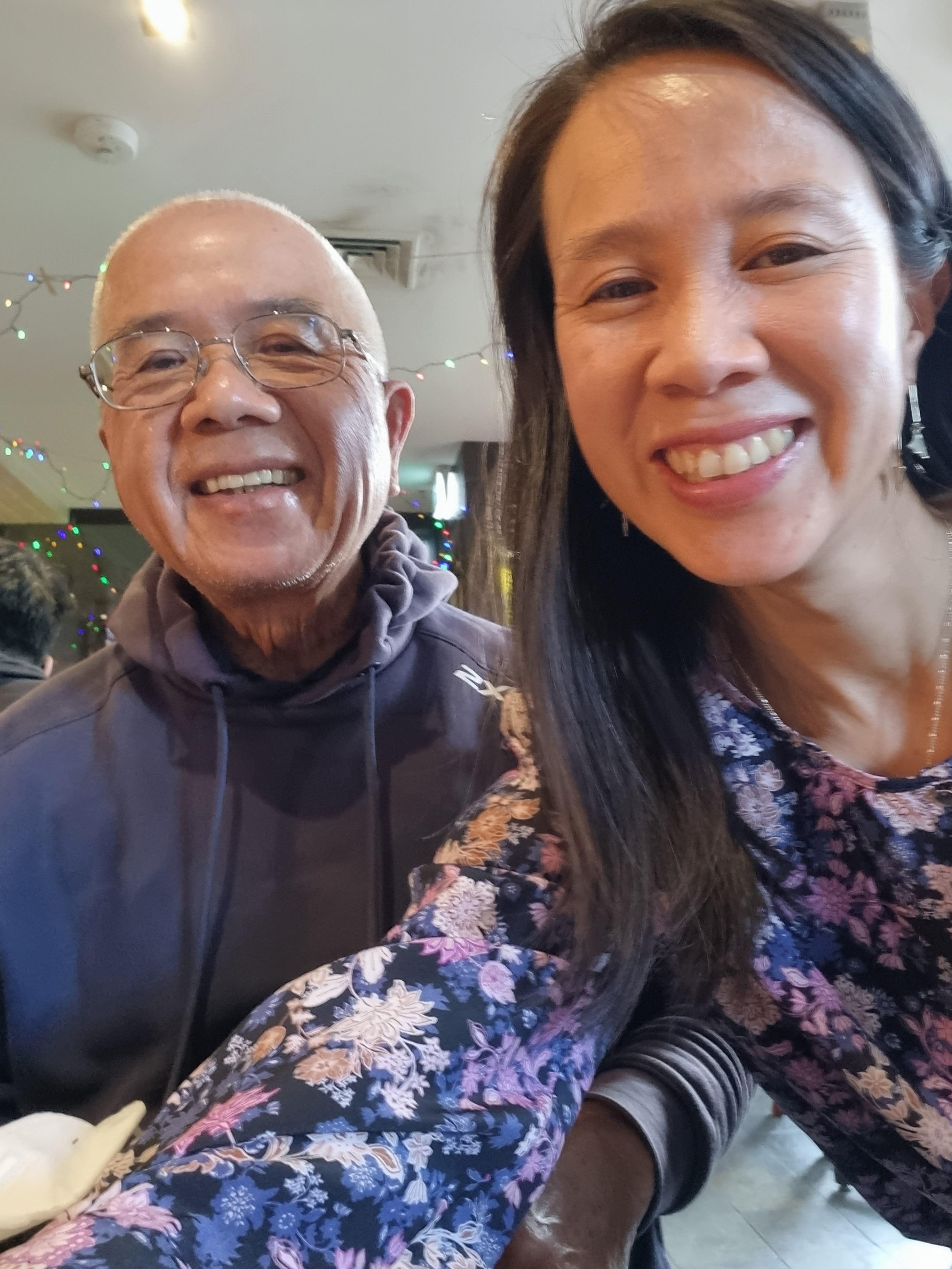 A selfie taken by a woman with black hair and earrings next to an older man in glasses and both of them are smiling.