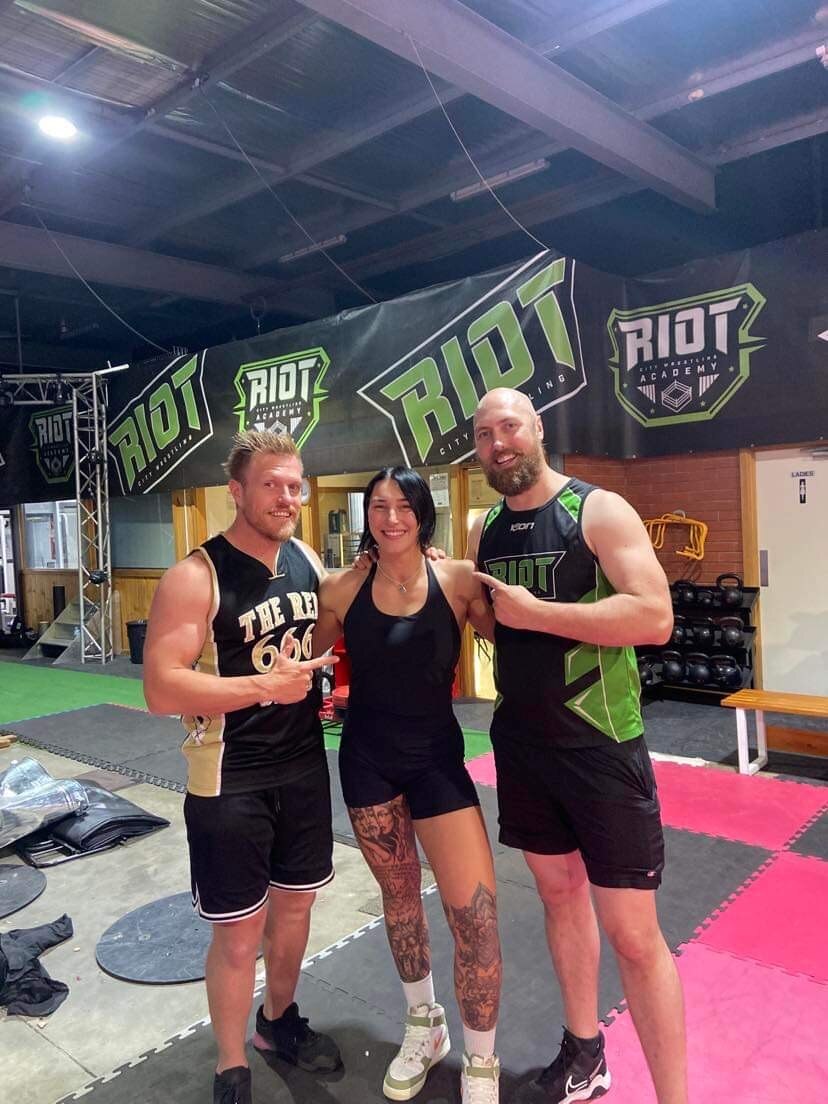 A muscly woman with short black hair wearing all black stands in between two muscly men in a wrestling gym.
