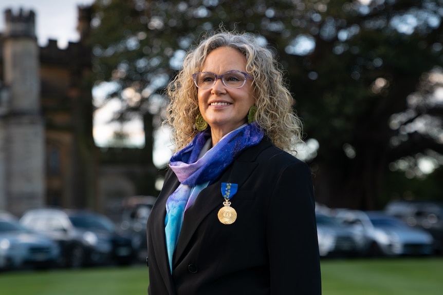 Leah Purcell smiles wearing glasses and a black blazer with a bright blue and purple scarf underneath. AM medal pinned to jacket