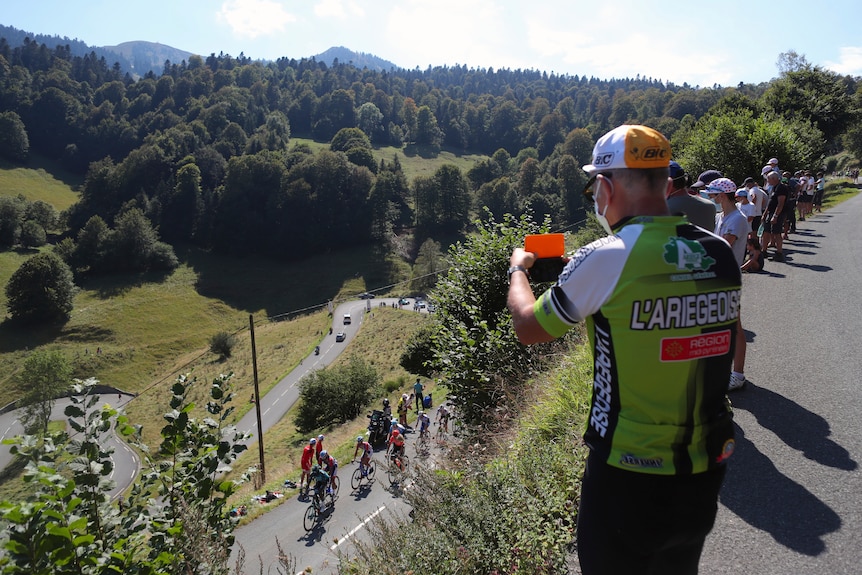 A man holds a camera overlooking bike riders below him on a road