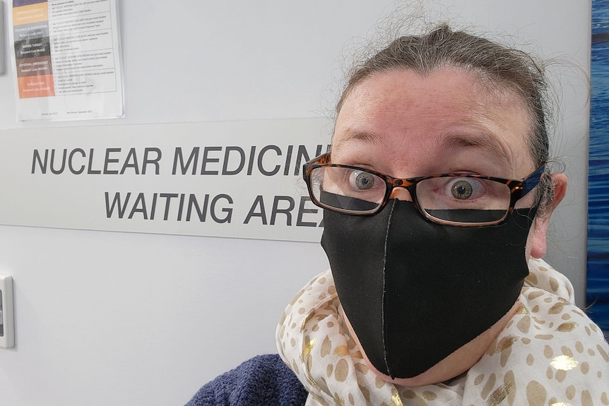 A close up of woman, brown hair tied back, glasses, wears a black mask, eyes wide, in front of Nuclear Medicine waiting area.