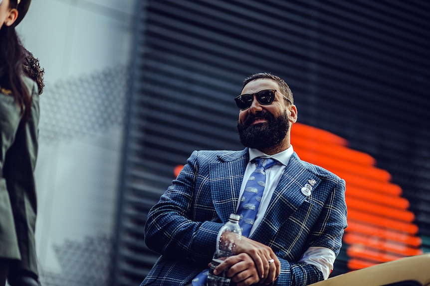 A man in a tweed jacket and dark sunglasses leans against a motorcycle.