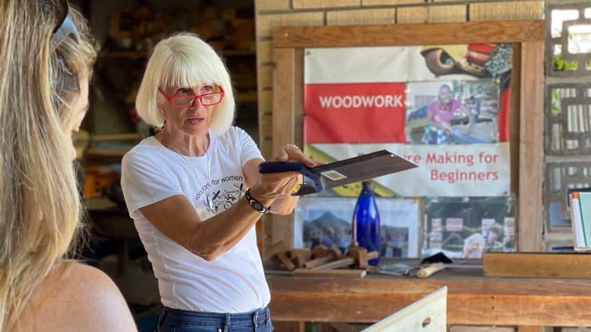A woman wearing jeans, a white t shirt and glasses holds up a hand saw.