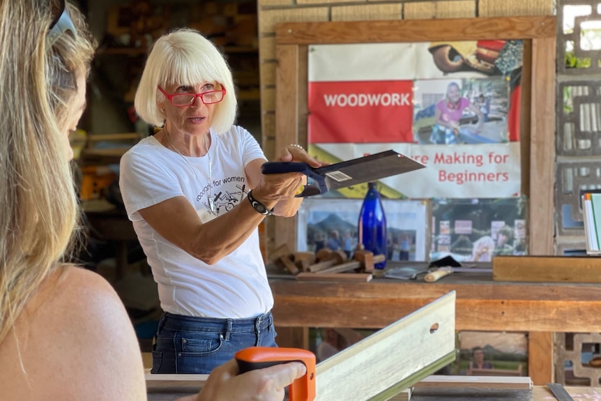 A woman wearing jeans, a white t shirt and glasses holds up a hand saw.