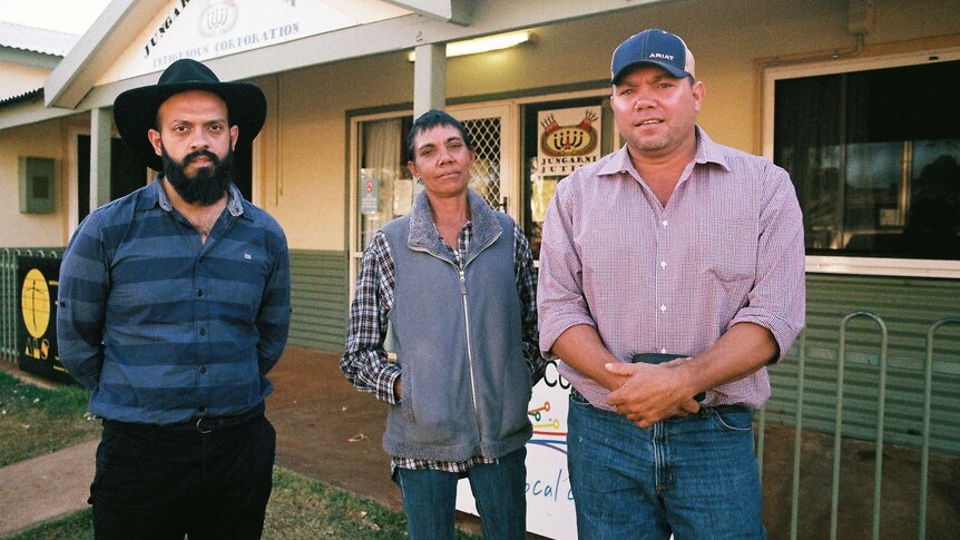 A woman and two men look at the camera while standing in front of a community hall.