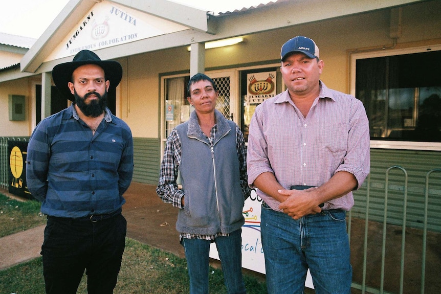 A woman and two men look at the camera while standing in front of a community hall.