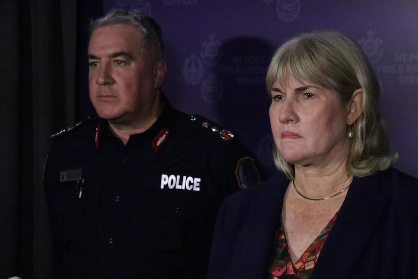 A woman stands at a press conference listening to questions from reporters, next to a policeman