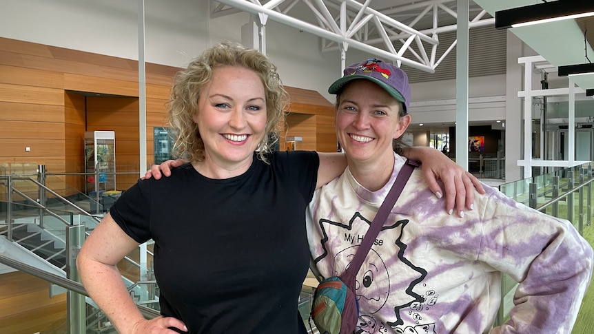 Zoe and Zan smile at the ABC in Melbourne. Zan wears a black tshirt and Zoe wears a purple tie-dye long sleeve and wears a cap.