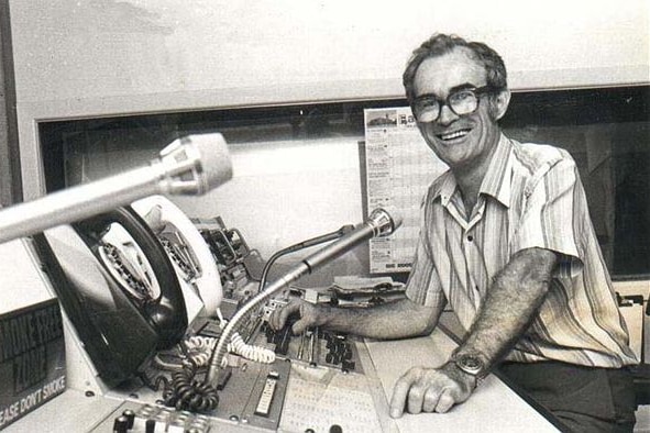 Black and white photo, Milton Moorhead sitting at a radio panel, microphones in shot, smiling. 