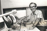 Black and white photo, Milton Moorhead sitting at a radio panel, microphones in shot, smiling. 
