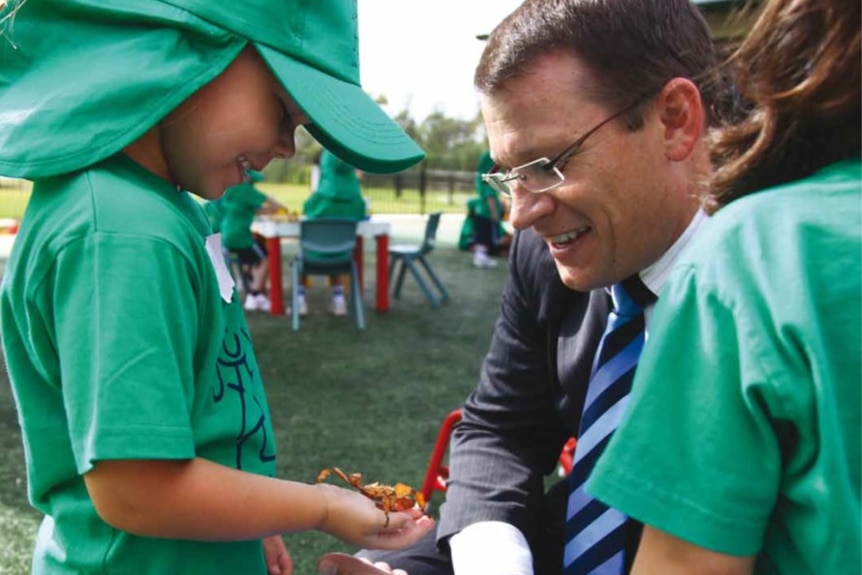 Paul Kidson wearing a suit, crouching, and looking at a stick insect a little boy is holding in his hand.