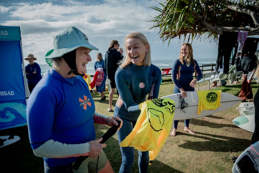 Two women, an older one on the left and younger one on the right, stand in wetsuits laughing at each other