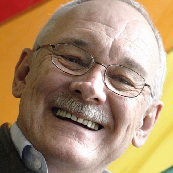 bald man with glasses smiling with rainbow flag in the background