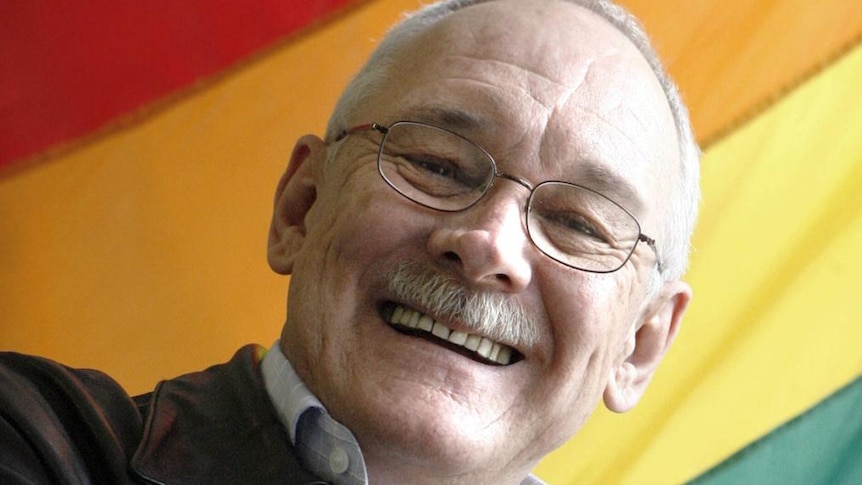 bald man with glasses smiling with rainbow flag in the background