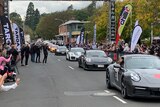 Cars are lined along a road as hundreds of people watch on and take photos. 
