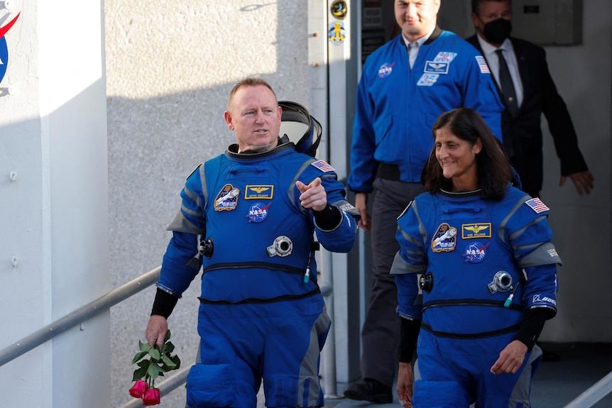 A man and a woman in bright blue spacesuits walk down a ramp or stairs looking happy.